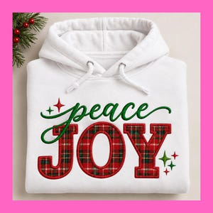 May include: A white hooded sweatshirt featuring the word peace in green cursive script above the word JOY in large, red and green plaid block letters. The design includes small red and green star accents on a plain white fabric background.
