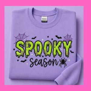 Peut inclure: Un sweat-shirt à col rond violet clair avec le texte SPOOKY en lettres vertes dégoulinantes et season en écriture noire. Le motif comprend des toiles d'araignée violettes, des chauves-souris noires, des étoiles et une araignée noire.