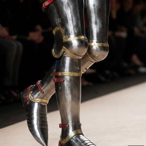 May include: Highly polished steel leg armour with gold detailing and red leather straps. The armour includes articulated knee and ankle sections, and a pointed toe design. The image highlights the craftsmanship of medieval-style armour.