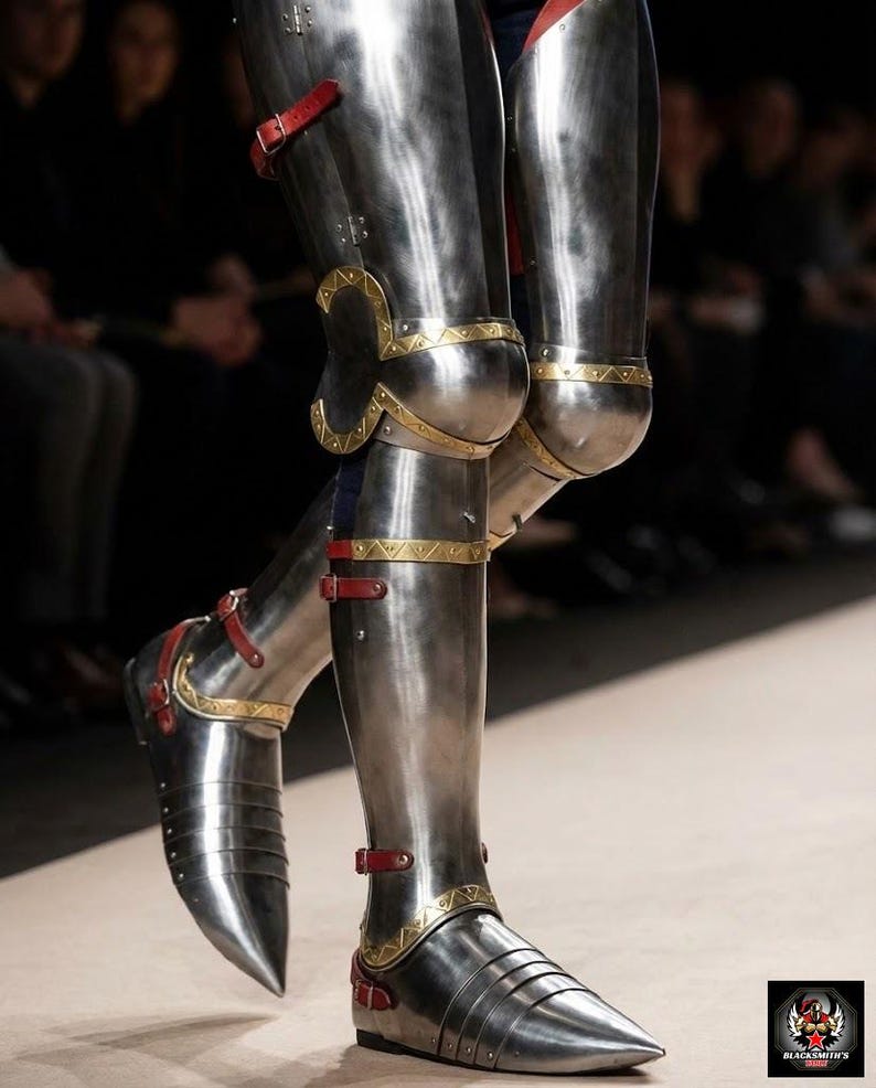 May include: Highly polished steel leg armour with gold detailing and red leather straps. The armour includes articulated knee and ankle sections, and a pointed toe design. The image highlights the craftsmanship of medieval-style armour.