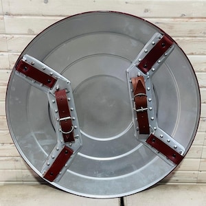 May include: A circular shield in silver with a red edge and leather straps. The shield has a metallic finish and is designed with riveted metal accents. The back features two brown leather straps for securing the shield. The shield is a replica of a superhero shield.