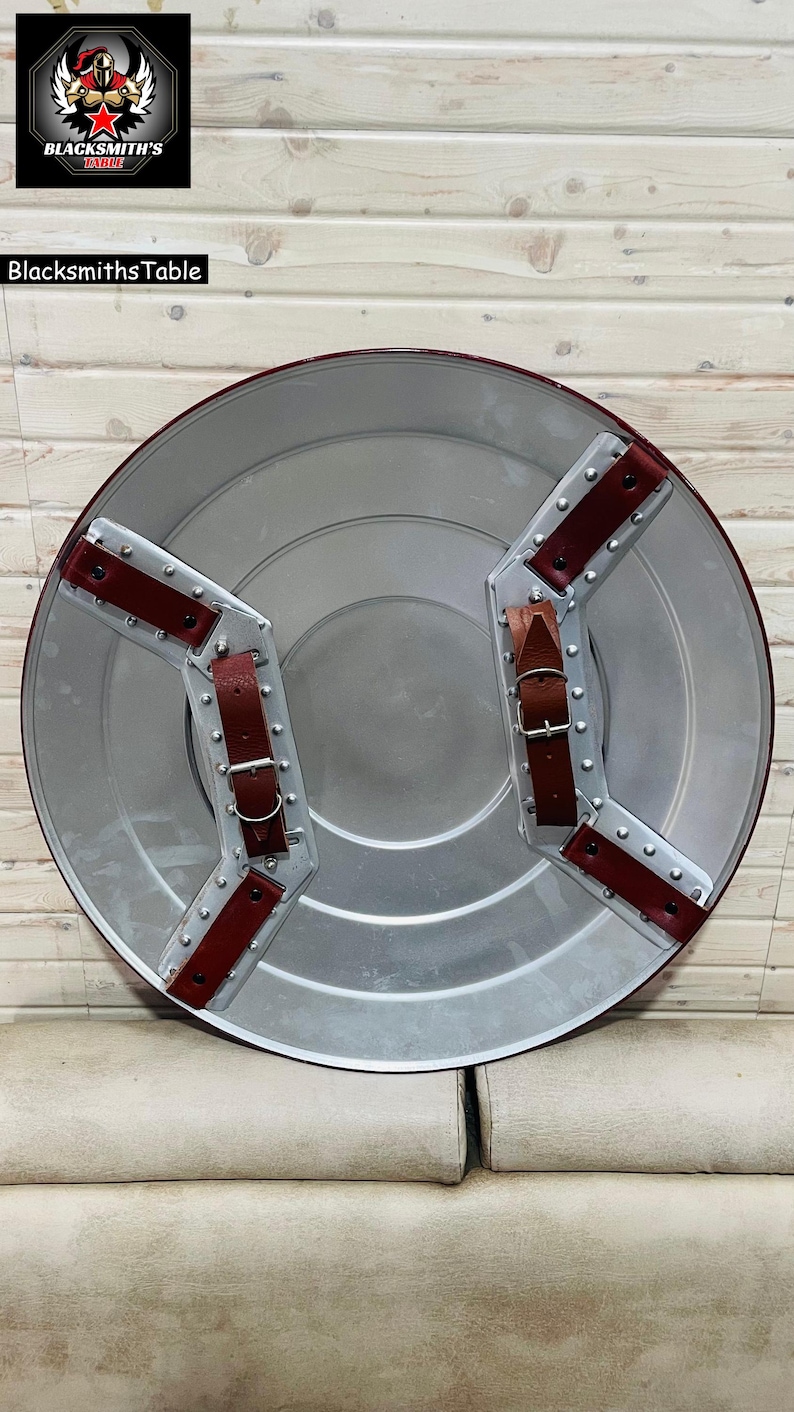 May include: A circular shield in silver with a red edge and leather straps. The shield has a metallic finish and is designed with riveted metal accents. The back features two brown leather straps for securing the shield. The shield is a replica of a superhero shield.