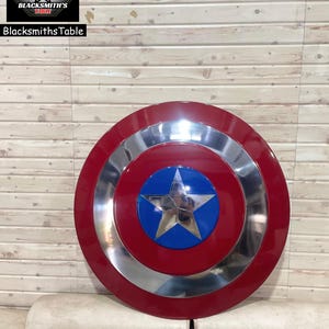 May include: A Captain America shield in red, silver, and blue. The shield has a central blue circle with a silver star, encircled by silver and red rings. It's set against a light brown wooden backdrop. The shield's diameter is approximately 76 cm.