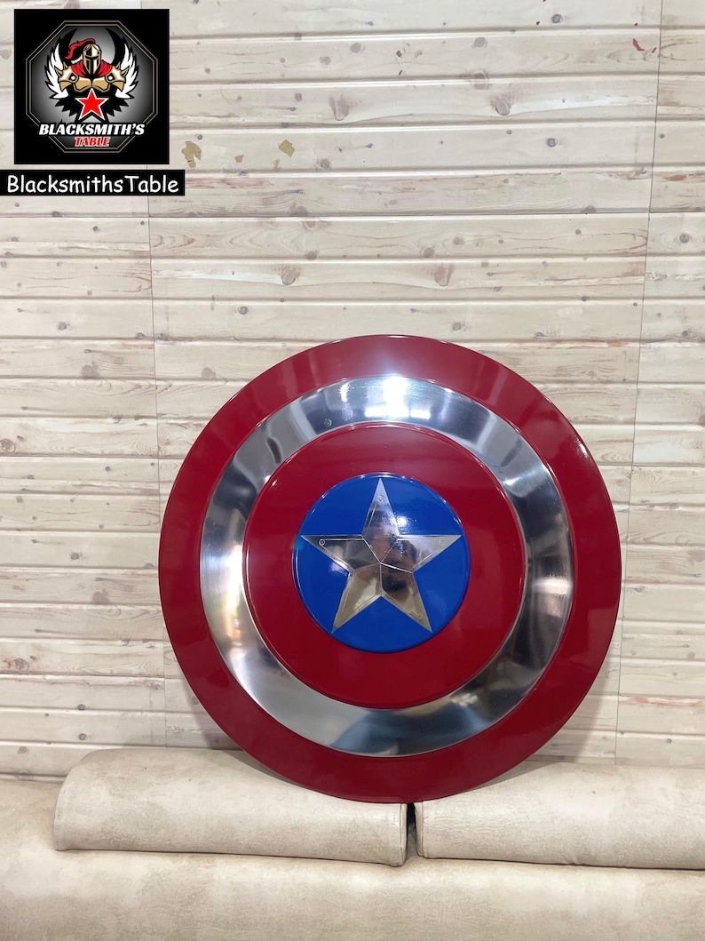 May include: A Captain America shield in red, silver, and blue. The shield has a central blue circle with a silver star, encircled by silver and red rings. It's set against a light brown wooden backdrop. The shield's diameter is approximately 76 cm.