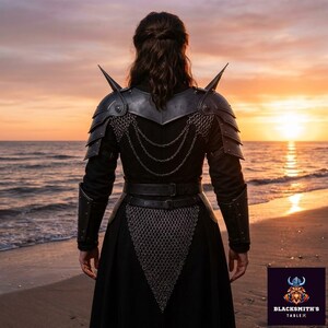 May include: A person wearing black armor with shoulder spikes, chainmail, and a belt, standing on a beach at sunset. The armor has a metallic appearance. The Blacksmith's Table logo is in the bottom right corner.