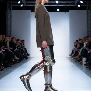 May include: A model on a runway wears a long-sleeved, olive-green top and metallic leg armour. The armour is silver with red straps and gold accents at the knees. The runway is white, and an audience is seated on either side.