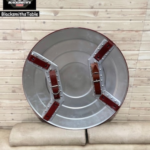 May include: A circular shield in silver, featuring red leather straps and rivets. The shield has a central, slightly raised area. The words "BlacksmithsTable" are visible in the upper left corner, along with a logo.