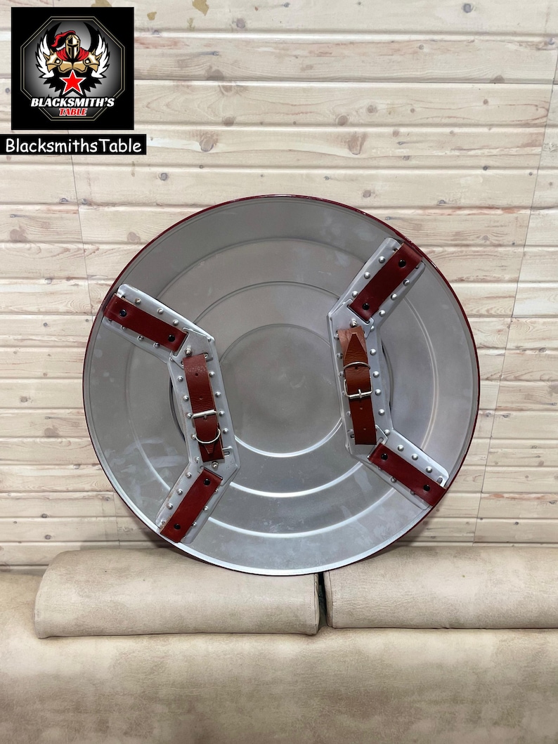 May include: A circular shield in silver, featuring red leather straps and rivets. The shield has a central, slightly raised area. The words "BlacksmithsTable" are visible in the upper left corner, along with a logo.