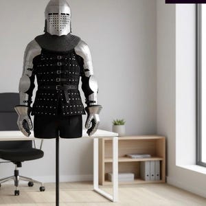 May include: A suit of medieval armour, including a helmet, chainmail, and metal plating, displayed on a black stand. The armour is black and silver, with a black tunic and shorts. The image is taken indoors, with a desk and shelving in the background.