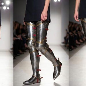 May include: Metallic silver leg armour with red and gold detailing, worn with a black top. The armour extends from the feet to the upper thighs. The image is taken on a runway with a blurred background.