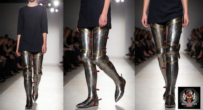 May include: Metallic silver leg armour with red and gold detailing, worn with a black top. The armour extends from the feet to the upper thighs. The image is taken on a runway with a blurred background.