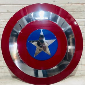 May include: A red, silver, and blue shield with a star emblem. The shield has a red outer ring, a silver middle ring, and a blue centre with a silver star. The shield is a replica of a superhero's shield.