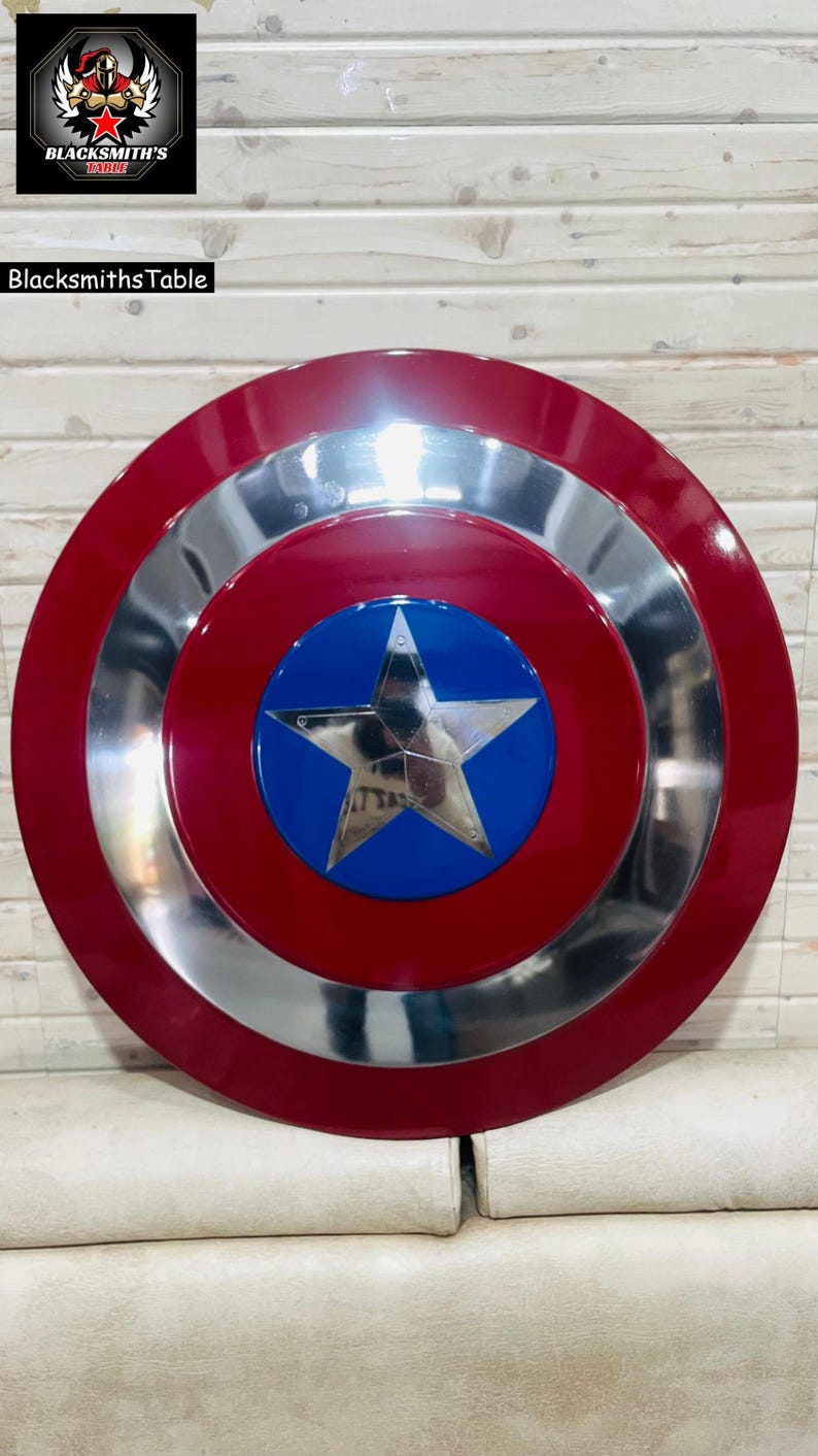 May include: A red, silver, and blue shield with a star emblem. The shield has a red outer ring, a silver middle ring, and a blue centre with a silver star. The shield is a replica of a superhero's shield.