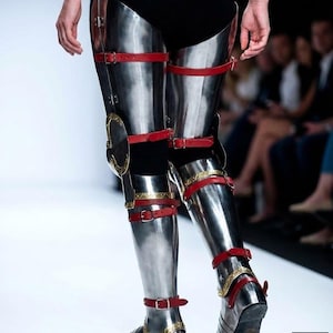 May include: Shiny metal leg armour with red leather straps and gold detailing. The armour is displayed on a catwalk, suggesting a medieval or fantasy theme. The design includes knee protection and foot coverings.