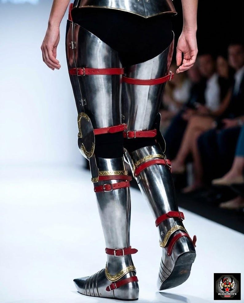 May include: Shiny metal leg armour with red leather straps and gold detailing. The armour is displayed on a catwalk, suggesting a medieval or fantasy theme. The design includes knee protection and foot coverings.