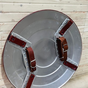 May include: A circular shield in silver with a red edge and brown leather straps. The shield has a riveted metal backing and two brown leather straps with buckles. The Blacksmith's Table logo is visible in the top left corner.