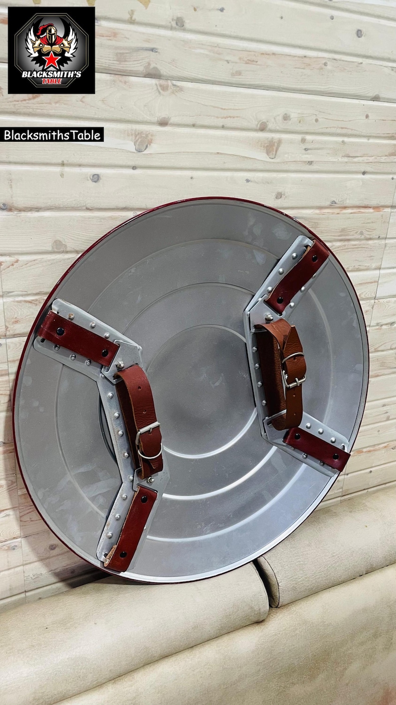 May include: A circular shield in silver with a red edge and brown leather straps. The shield has a riveted metal backing and two brown leather straps with buckles. The Blacksmith's Table logo is visible in the top left corner.