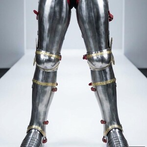 May include: Highly polished steel leg armour with red leather and gold-coloured trim. The armour features articulated plates and straps for fastening. The design is reminiscent of medieval knight armour.