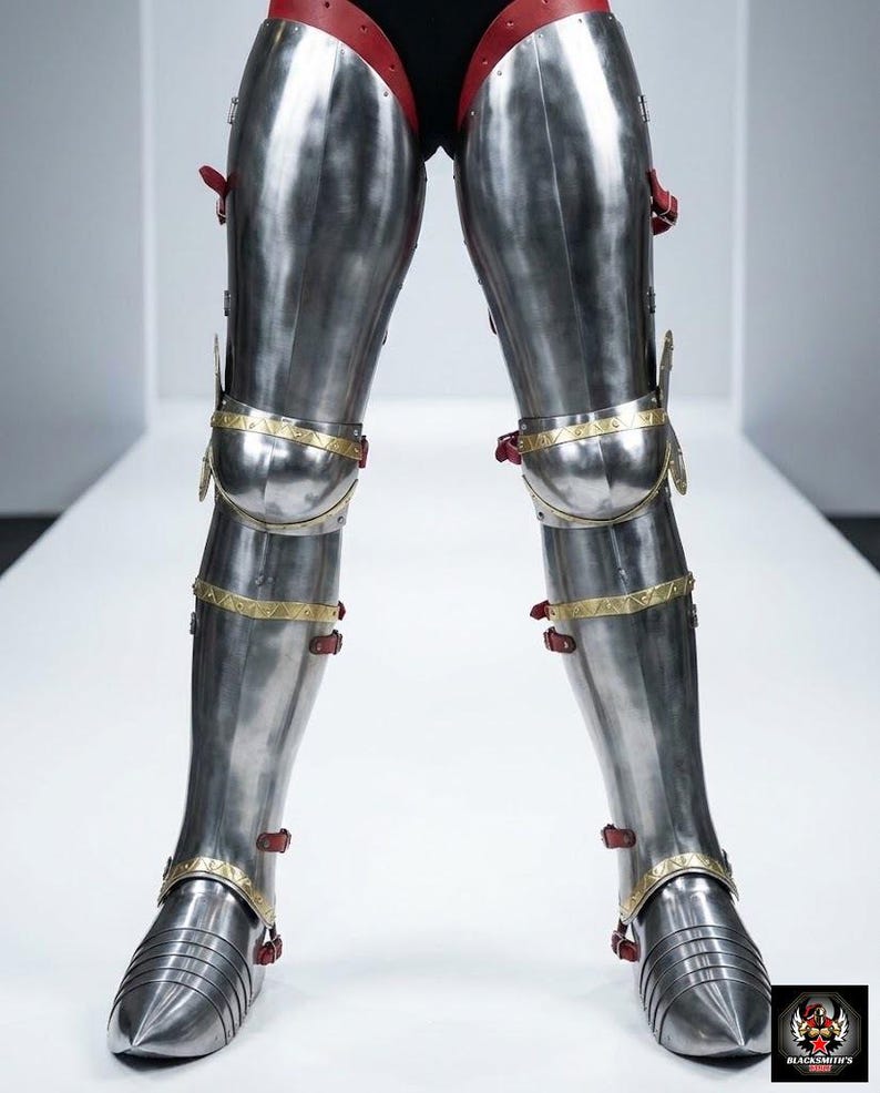 May include: Highly polished steel leg armour with red leather and gold-coloured trim. The armour features articulated plates and straps for fastening. The design is reminiscent of medieval knight armour.