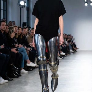 May include: A model on a catwalk wearing a black t-shirt and polished metal leg armour. The armour includes knee and ankle protection, with decorative gold-coloured accents. The catwalk is in a well-lit space with an audience.