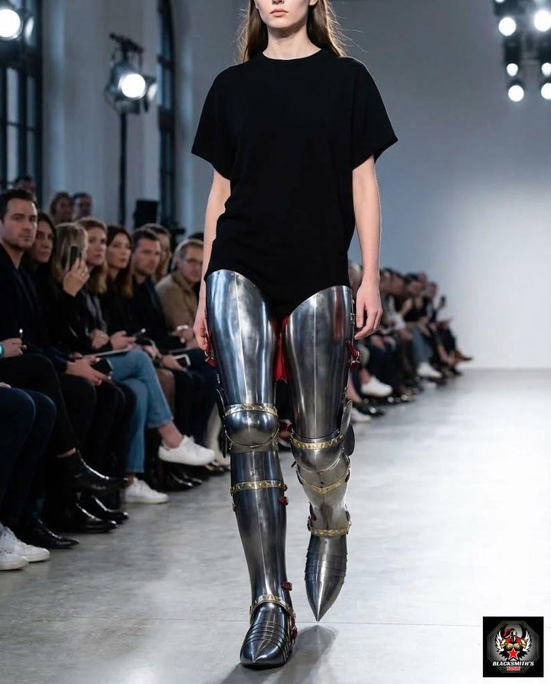 May include: A model on a catwalk wearing a black t-shirt and polished metal leg armour. The armour includes knee and ankle protection, with decorative gold-coloured accents. The catwalk is in a well-lit space with an audience.