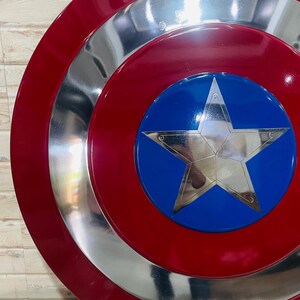 May include: A circular shield in red, silver, and blue, featuring a large silver star at its centre. The shield has a red outer ring, a silver middle ring, and a blue central disc. A replica of a superhero's shield.