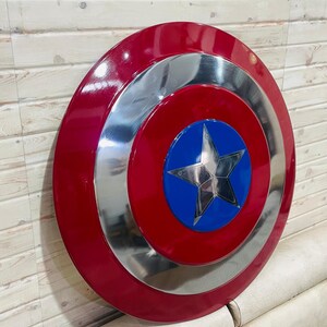 May include: A red, silver, and blue shield with a star emblem. The shield has a glossy finish and a circular design. The shield is a prop or collectible item. The text "Blacksmiths Table" is visible in the image.