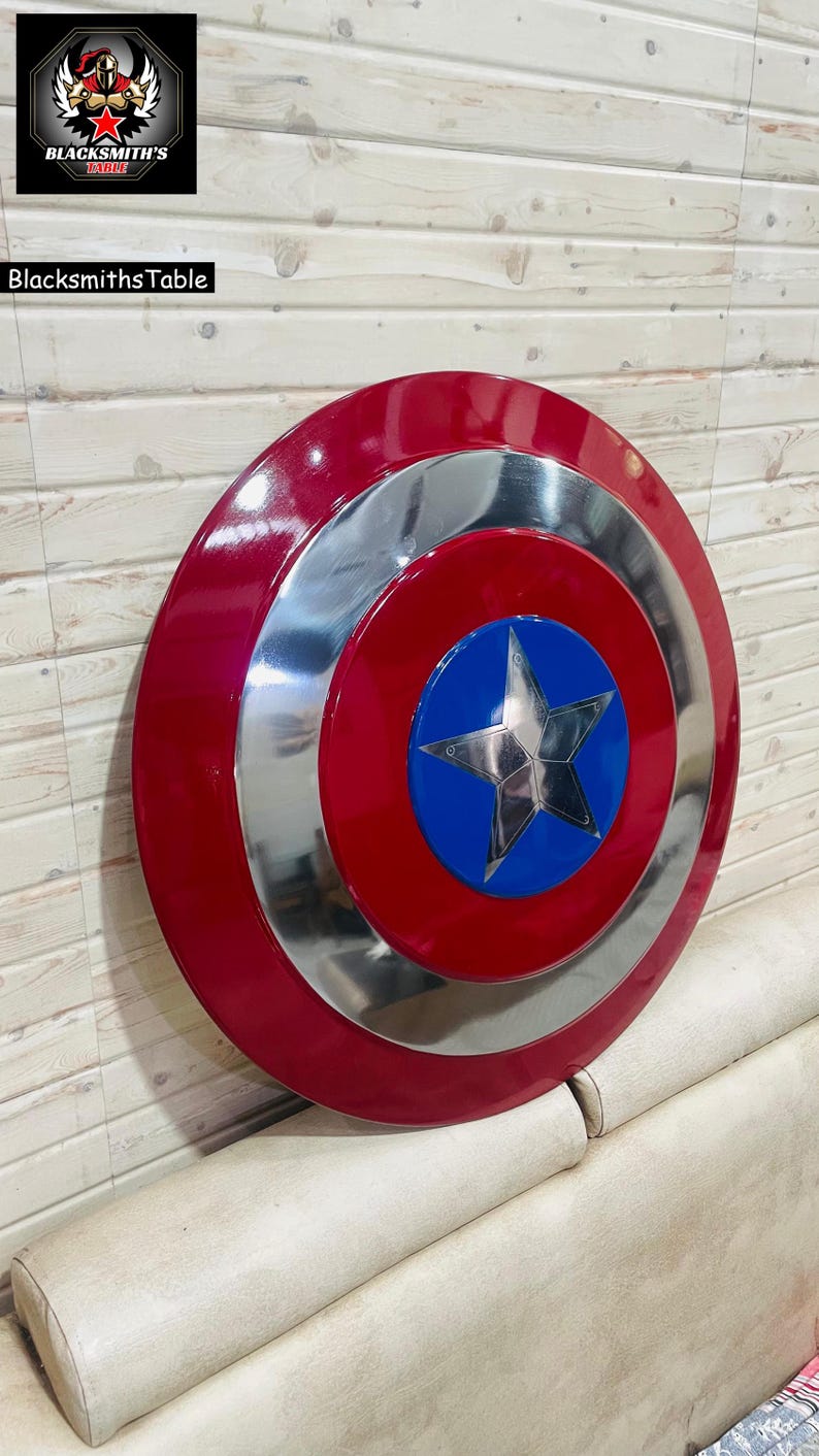 May include: A red, silver, and blue shield with a star emblem. The shield has a glossy finish and a circular design. The shield is a prop or collectible item. The text "Blacksmiths Table" is visible in the image.