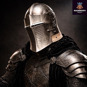 Puede incluir: Imagen detallada de una armadura medieval, que incluye un casco plateado con visera perforada y detalles intrincados. La armadura presenta un acabado oscuro y texturizado, con una capa negra sobre los hombros. Fondo oscuro.