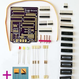 May include: A purple circuit board with the text "AYAB-Shield V1.4TH" and "KH 910/950/i/e". The board is surrounded by various electronic components, including LEDs, resistors, capacitors, and a fuse.