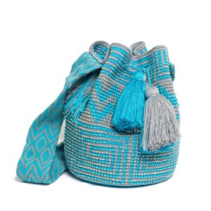 Puede incluir: Un bolso de cubo tejido turquesa y gris con un patrón geométrico. El bolso presenta una correa larga con un diseño de diamantes y está adornado con borlas turquesas y grises. El bolso mide aproximadamente 30 cm de alto.