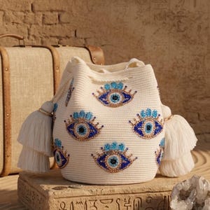 Puede incluir: Un bolso tipo saco de color crema con un patr&oacute;n repetitivo de motivos de ojos turcos hechos con cuentas en tonos azul, dorado y blanco. El bolso tiene un cierre de cord&oacute;n y est&aacute; decorado con grandes borlas blancas en capas a ambos lados.