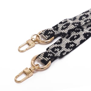 May include: Close-up of a black and silver leopard print bag strap with gold-colored metal clasps. The strap is made of a woven material with a repeating animal print pattern. The clasps are attached to the strap with gold-colored rings.