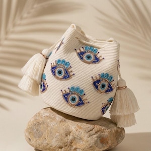 Puede incluir: Un bolso tipo cubo de color crema con textura tejida y m&uacute;ltiples motivos bordados de ojos turcos en tonos azul, dorado y negro. El bolso incluye un cierre de cord&oacute;n y dos grandes borlas decorativas de color crema en los laterales.