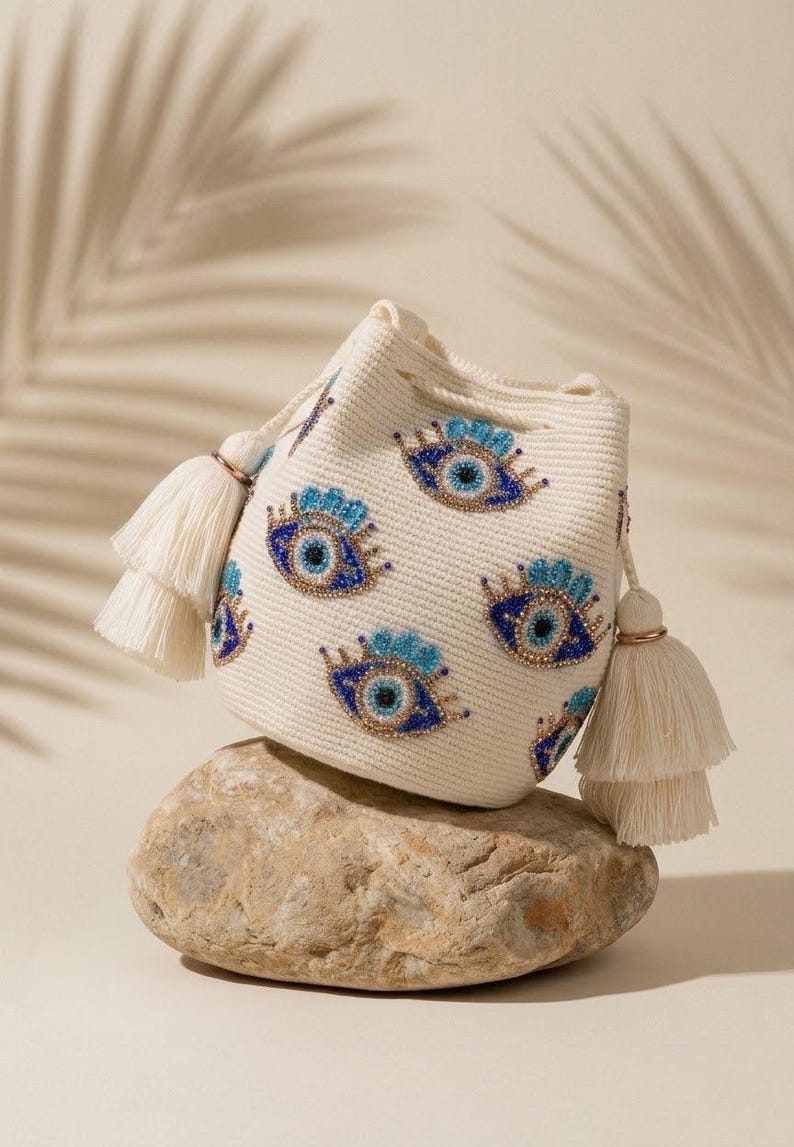 Puede incluir: Un bolso tipo cubo de color crema con textura tejida y m&uacute;ltiples motivos bordados de ojos turcos en tonos azul, dorado y negro. El bolso incluye un cierre de cord&oacute;n y dos grandes borlas decorativas de color crema en los laterales.