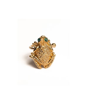 Puede incluir: Un anillo de rana dorado con detalles texturizados. La rana tiene ojos de piedra verde y está engastada en una fina banda dorada. Una pieza de joyería caprichosa.
