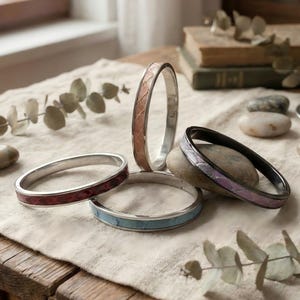 May include: A selection of silver bangles with patterned inlays in various colours. The bangles are in shades of red, blue, pink, and purple. The jewellery is displayed on a neutral cloth on a wooden table.
