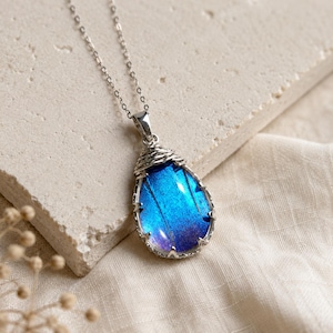 May include: A teardrop-shaped pendant necklace featuring a vibrant iridescent blue butterfly wing encased in a silver-toned metal frame with wire-wrapped detailing at the top, suspended from a delicate silver-toned chain link necklace.