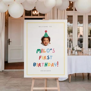 Könnte beinhalten: Ein weißes Schild auf einer Staffelei mit dem Text "WELCOME TO" und "MILO'S FIRST BIRTHDAY!" in bunten Buchstaben. Ein Foto eines lächelnden Babys mit Partyhut befindet sich über dem Text. Das Datum "08.20.20XX" steht unten.