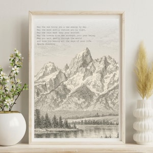 May include: A framed print with a pencil sketch of mountains and a lake. The artwork includes the text "May the sun bring you a new energy by day...Apache Blessing." The print is displayed on a white shelf with a vase of flowers and a decorative vase.