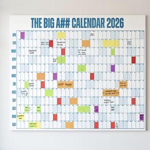 Könnte beinhalten: Ein großer weißer Kalender für das Jahr 2026 mit dem Text "THE BIG A## CALENDAR 2026" in Blau. Das Kalendarium ist mit bunten Haftnotizen und handschriftlichen Notizen gefüllt, die verschiedene Ereignisse markieren.