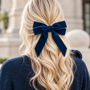 May include: A deep navy blue velvet hair bow with long, flowing tails. The bow is constructed from a soft, plush fabric with a subtle sheen, featuring a central knot detail that secures the loops and tails in a classic, symmetrical arrangement.