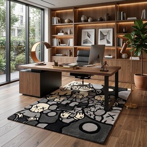 May include: A rectangular area rug featuring a repeating graphic pattern of cartoon-style figures with crossed-out eyes in shades of gray, black, and cream. The rug is positioned on a wooden floor beneath a wooden desk with a computer and lamp.