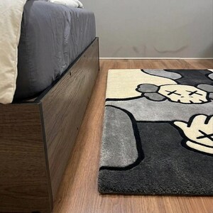 K&ouml;nnte beinhalten: Ein rechteckiger Teppich mit einem stilisierten Grafikdesign, das einen totenkopfartigen Charakterkopf und eine Hand mit vier Fingern zeigt. Der Teppich besteht aus cremefarbenen, hellgrauen und dunkel anthrazitfarbenen Bereichen mit schwarzen Konturen.
