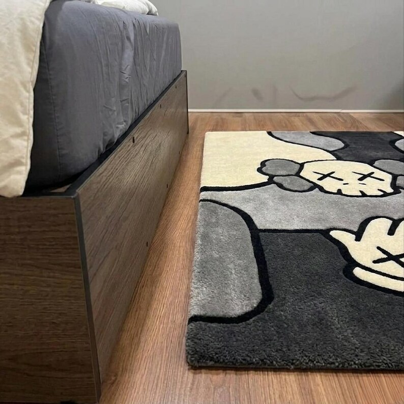 K&ouml;nnte beinhalten: Ein rechteckiger Teppich mit einem stilisierten Grafikdesign, das einen totenkopfartigen Charakterkopf und eine Hand mit vier Fingern zeigt. Der Teppich besteht aus cremefarbenen, hellgrauen und dunkel anthrazitfarbenen Bereichen mit schwarzen Konturen.