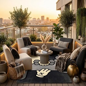 K&ouml;nnte beinhalten: Ein Balkon-Sitzbereich mit zwei cremefarbenen Sesseln, zwei dunkelgrauen Sesseln und einem runden, geriffelten Tisch in der Mitte. Ein gro&szlig;er Teppich zeigt eine stilisierte Cartoon-Handgrafik. Olivenb&auml;ume in T&ouml;pfen und ein Korb sind vorhanden.