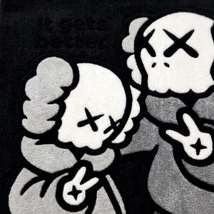 K&ouml;nnte beinhalten: Ein getufteter Teppich in Schwarz, Wei&szlig; und Grau mit zwei cartoonartigen Figuren, die X-f&ouml;rmige Augen und Friedenszeichen an den H&auml;nden haben. Der Text 'it gets better' ist in der oberen linken Ecke auf dunklem Grund eingestickt.