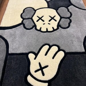 K&ouml;nnte beinhalten: Ein getufteter Teppich mit einem stilisierten Totenkopf-Charakter mit X-f&ouml;rmigen Augen und wolkenartigen Ohren, positioniert &uuml;ber einer Hand im Cartoon-Stil mit einem X-Zeichen auf der Handfl&auml;che. Das Design verwendet Creme, Hellgrau und Dunkelgrau.