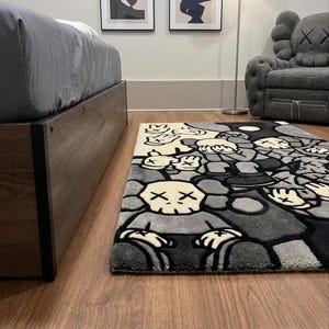 May include: A rectangular area rug featuring a repeating graphic pattern of cartoon-style characters with skull-like heads and X-shaped eyes. The design uses a monochromatic color palette of black, gray, and cream, with multiple hands shown in various poses.