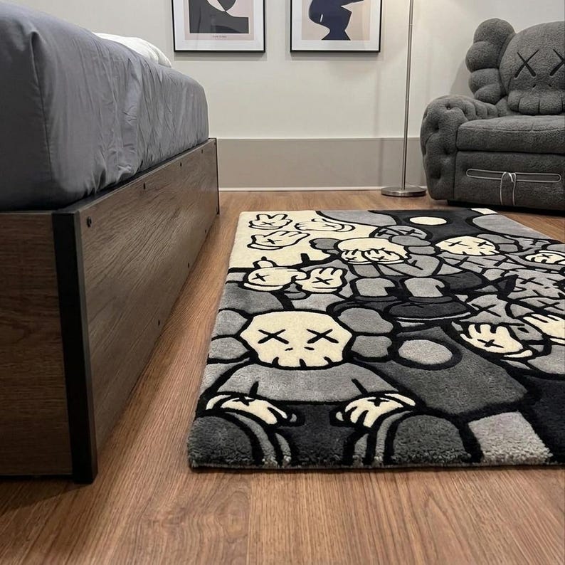 May include: A rectangular area rug featuring a repeating graphic pattern of cartoon-style characters with skull-like heads and X-shaped eyes. The design uses a monochromatic color palette of black, gray, and cream, with multiple hands shown in various poses.